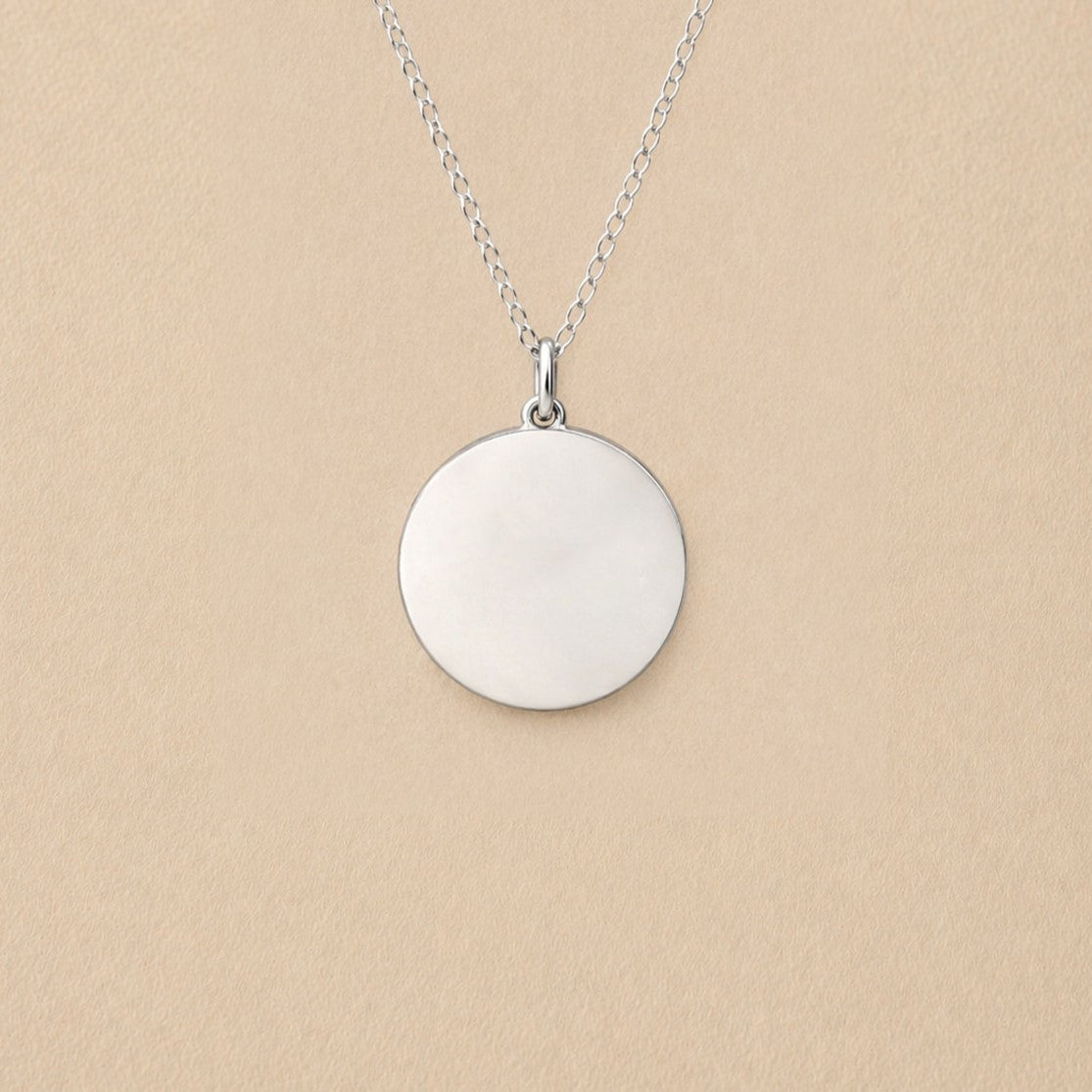 Collier "Reflet" avec médaille ronde personnalisé en acier inoxydable argenté, posé sur un fond beige texturé