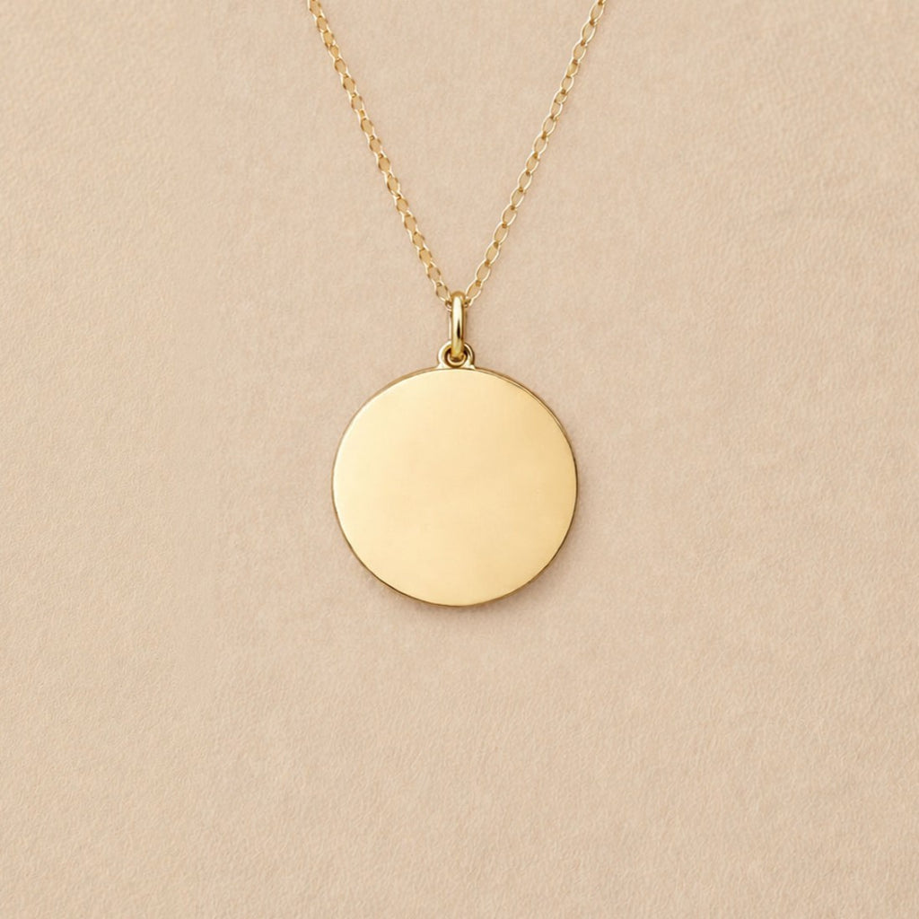 Collier "Reflet" avec médaille ronde personnalisé en acier inoxydable doré, posé sur un fond beige texturé