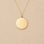 Collier "Reflet" avec médaille ronde personnalisé en acier inoxydable doré, posé sur un fond beige texturé