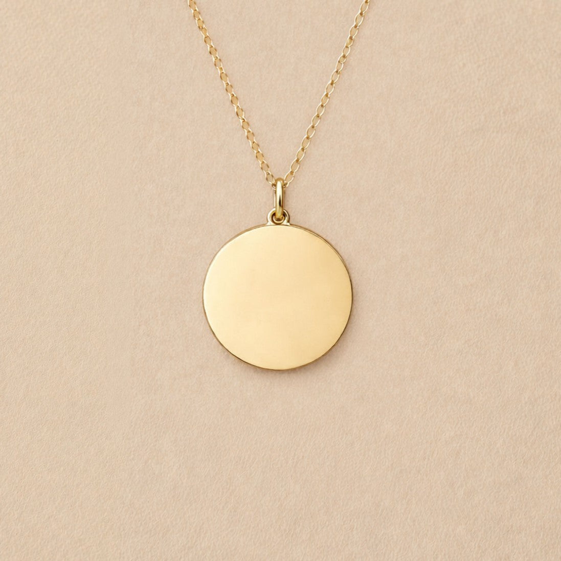 Collier "Reflet" avec médaille ronde personnalisé en acier inoxydable doré, posé sur un fond beige texturé