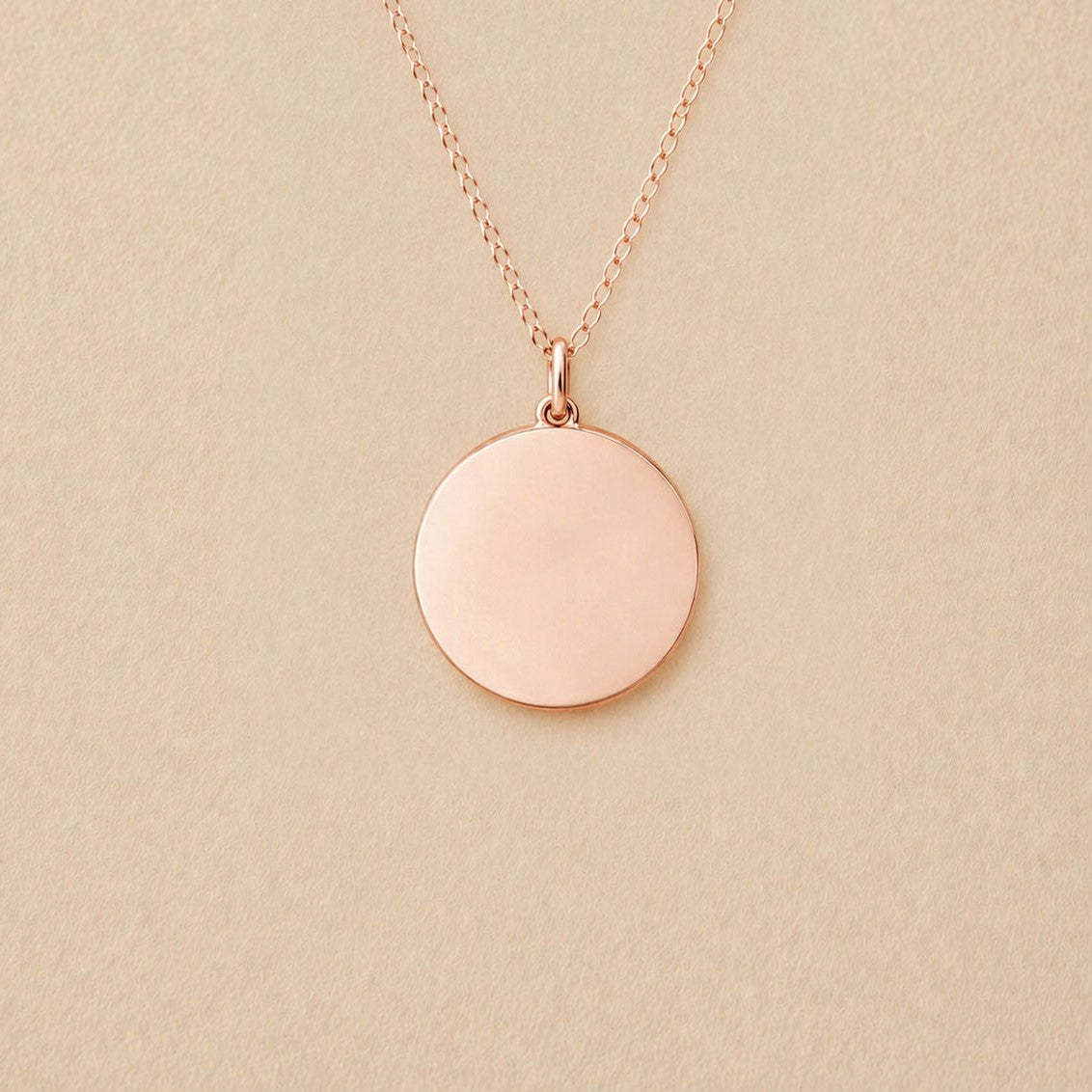 Collier "Reflet" avec médaille ronde personnalisé en acier inoxydable couleur or rose, posé sur un fond beige texturé