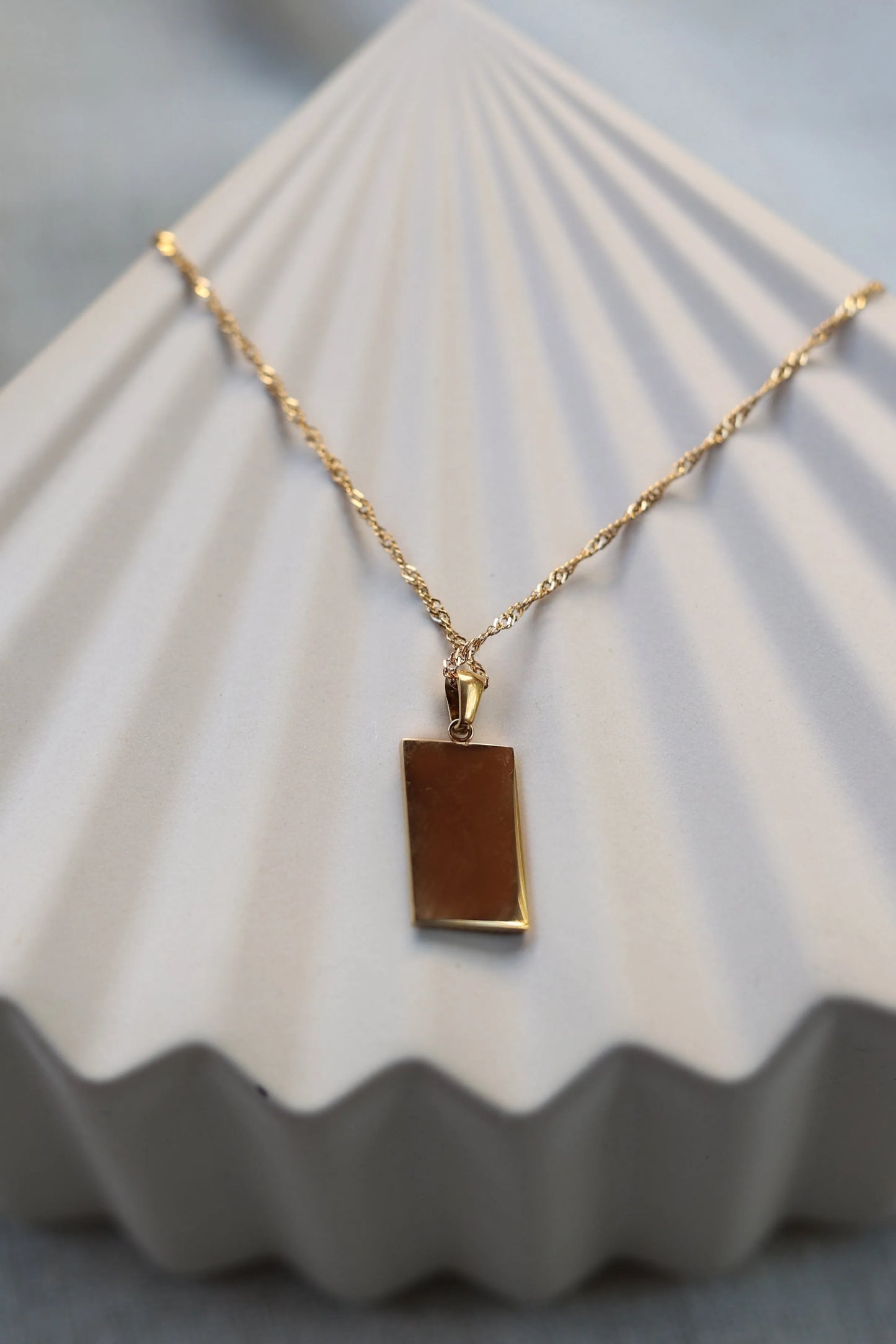 collier Résilience en acier doré avec pendentif rectangulaire gravé de rayons, personnalisable au verso, présenté côté verso sur fond beige texturé.