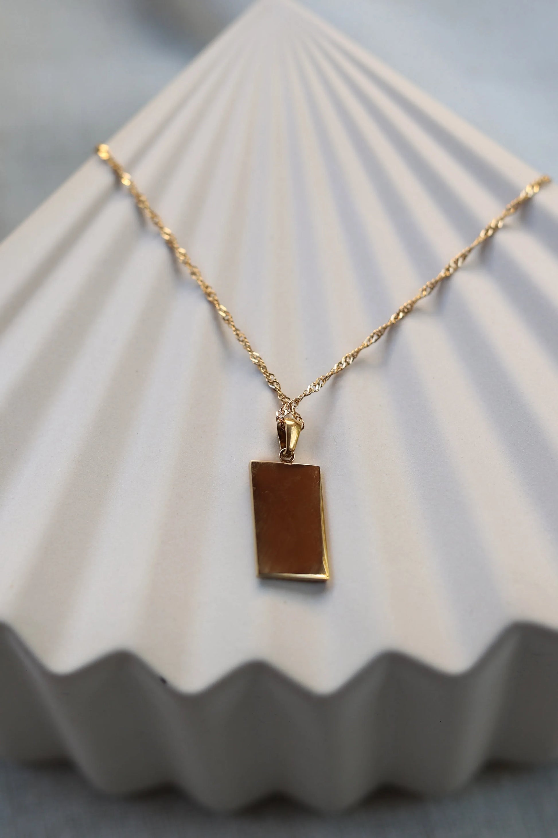 collier Résilience en acier doré avec pendentif rectangulaire gravé de rayons, personnalisable au verso, présenté côté verso sur fond beige texturé.