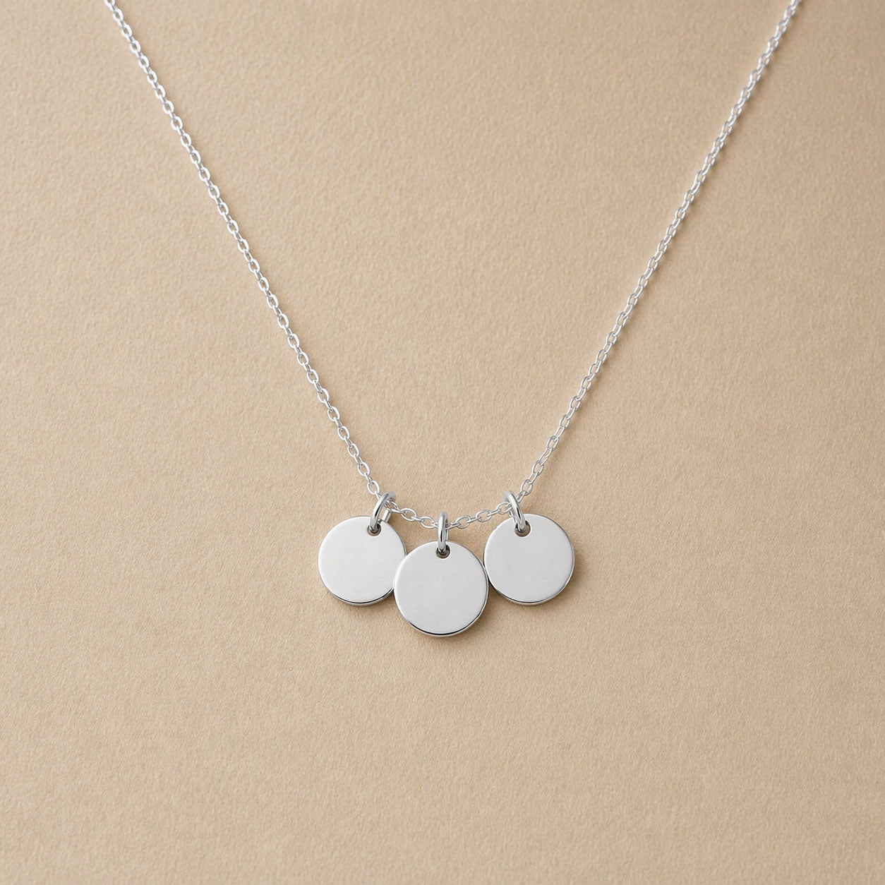 Collier "Inspiration" en acier inoxydable argenté, composé de trois médaillons personnalisables, présenté sur un fond beige texturé.
