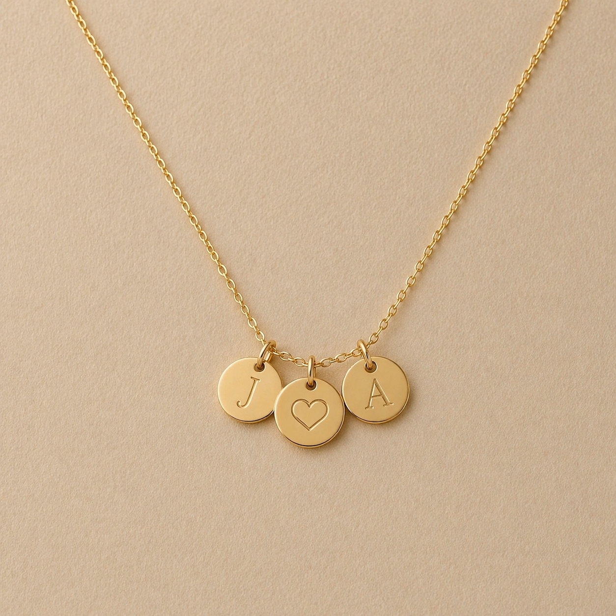 Collier "Inspiration" en acier inoxydable doré, composé de trois médaillons personnalisés avec J, coeur et A, présenté sur un fond beige texturé.