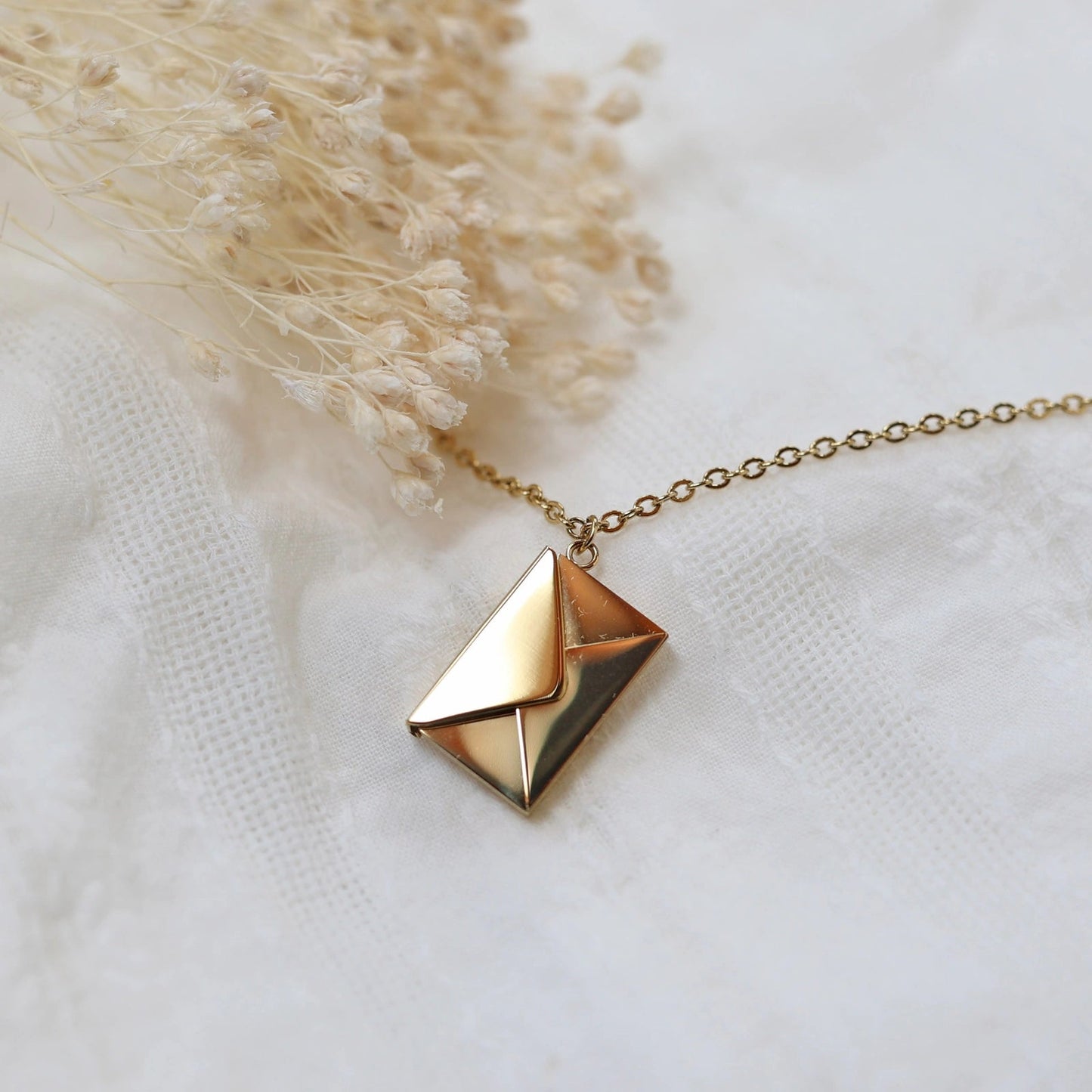 Collier "Nid secret", pendentif en forme d'enveloppe dorée fermée, posé avec soin sur un fond beige texturé.