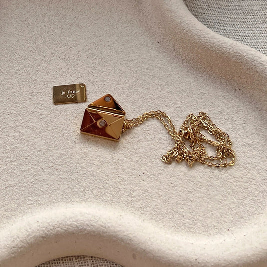 Collier doré en acier inoxydable en forme d'enveloppe ouverte, laissant apparaître un mot tendre, déposé avec soin sur un support beige.
