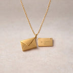 Collier "Nid secret", pendentif en forme d'enveloppe dorée ouverte, dévoilant un message "je t'aime", posé avec soin sur un fond beige texturé