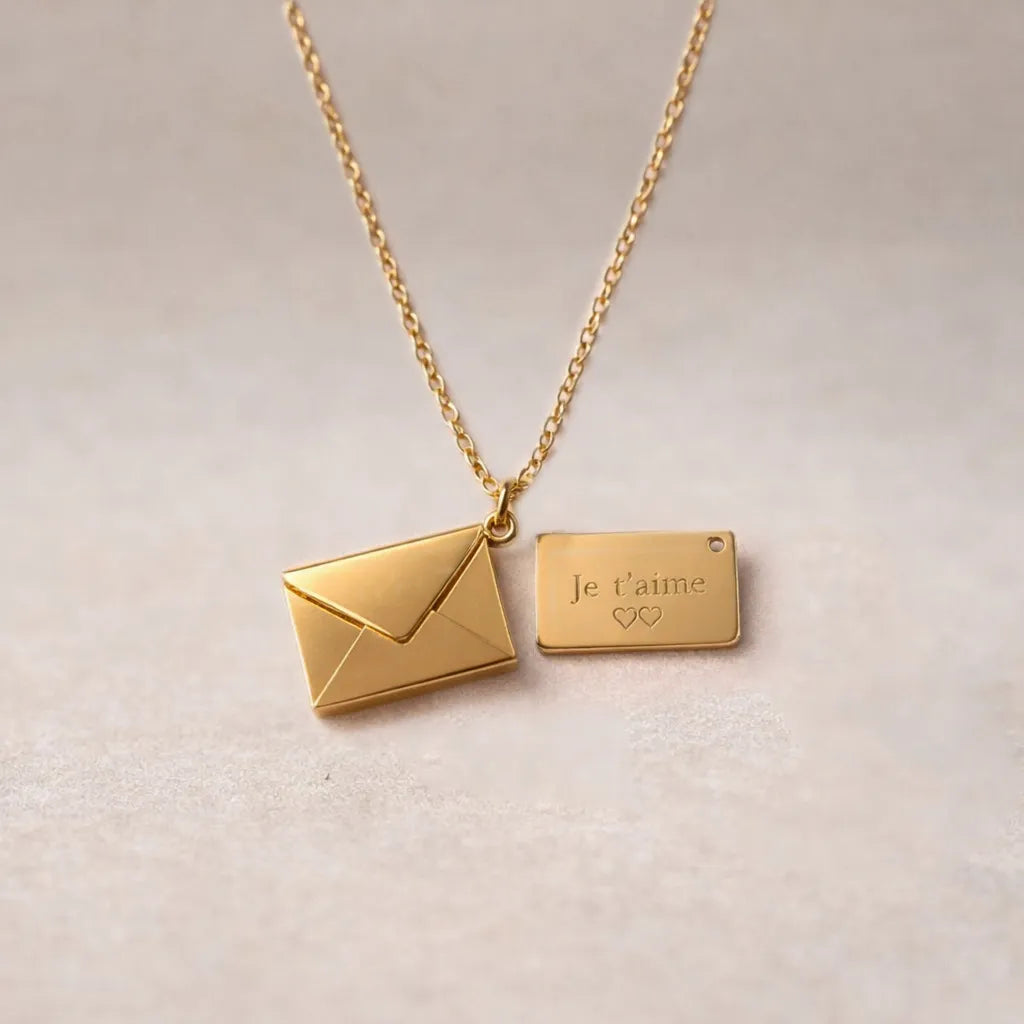 Collier "Nid secret", pendentif en forme d'enveloppe dorée ouverte, dévoilant un message "je t'aime", posé avec soin sur un fond beige texturé