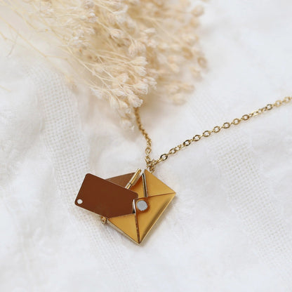 Collier "Nid secret", pendentif en forme d'enveloppe dorée ouverte, dévoilant un message, posé avec soin sur un fond beige texturé.