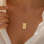 Collier Tracé en acier inoxydable doré, avec médaille rectangulaire gravé du prénom de Laura, porté autour du cou. Le modèle porte une chemise blanche chic.