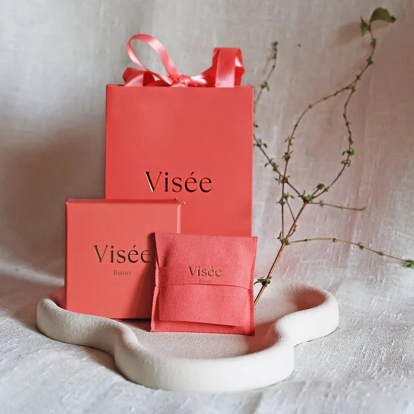 Vue d'ensemble de l'écrin, sachet et coffrets corail Visée Bijoux , délicatement posés sur un socle beige.