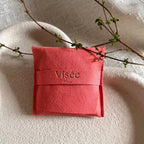 Pochon cadeau en suédine corail Visée Bijoux, posé sur un tissu beige plissé avec une branche végétale en diagonale.