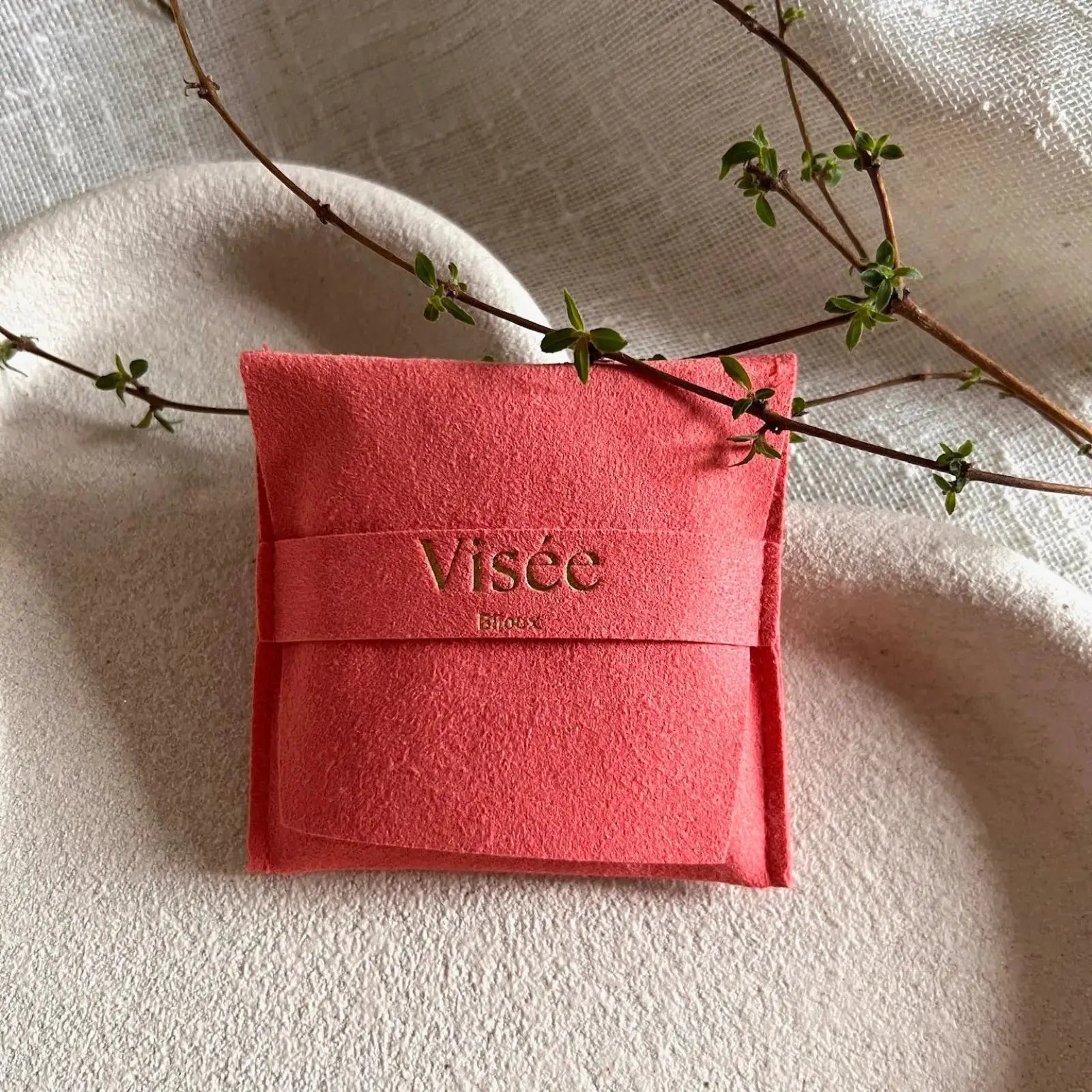Pochon cadeau en suédine corail Visée Bijoux, posé sur un tissu beige plissé avec une branche végétale en diagonale.