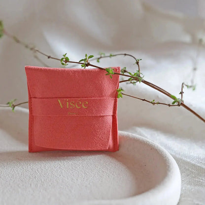 Pochon cadeau en suédine corail Visée Bijoux, posé sur un tissu beige plissé avec une branche végétale en diagonale.