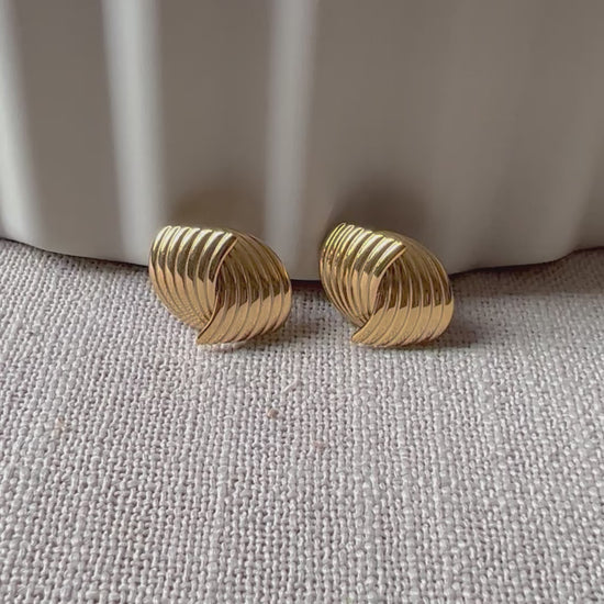 Vidéo des boucles d’oreilles torsadées Métamorphose en acier inoxydable doré, posées sur un tissu beige texturé.
