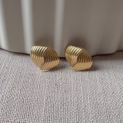 Vidéo des boucles d’oreilles torsadées Métamorphose en acier inoxydable doré, posées sur un tissu beige texturé.