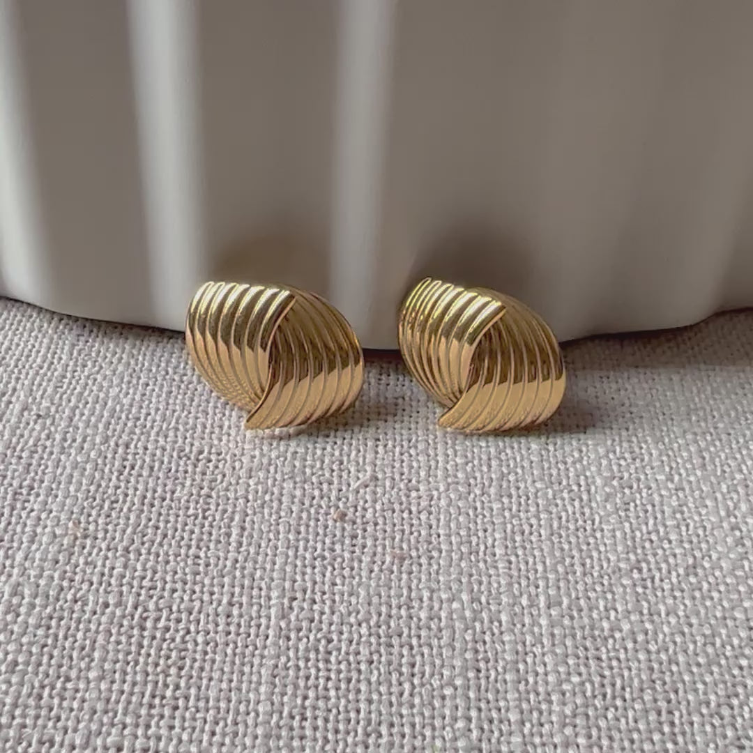 Vidéo des boucles d’oreilles torsadées Métamorphose en acier inoxydable doré, posées sur un tissu beige texturé.