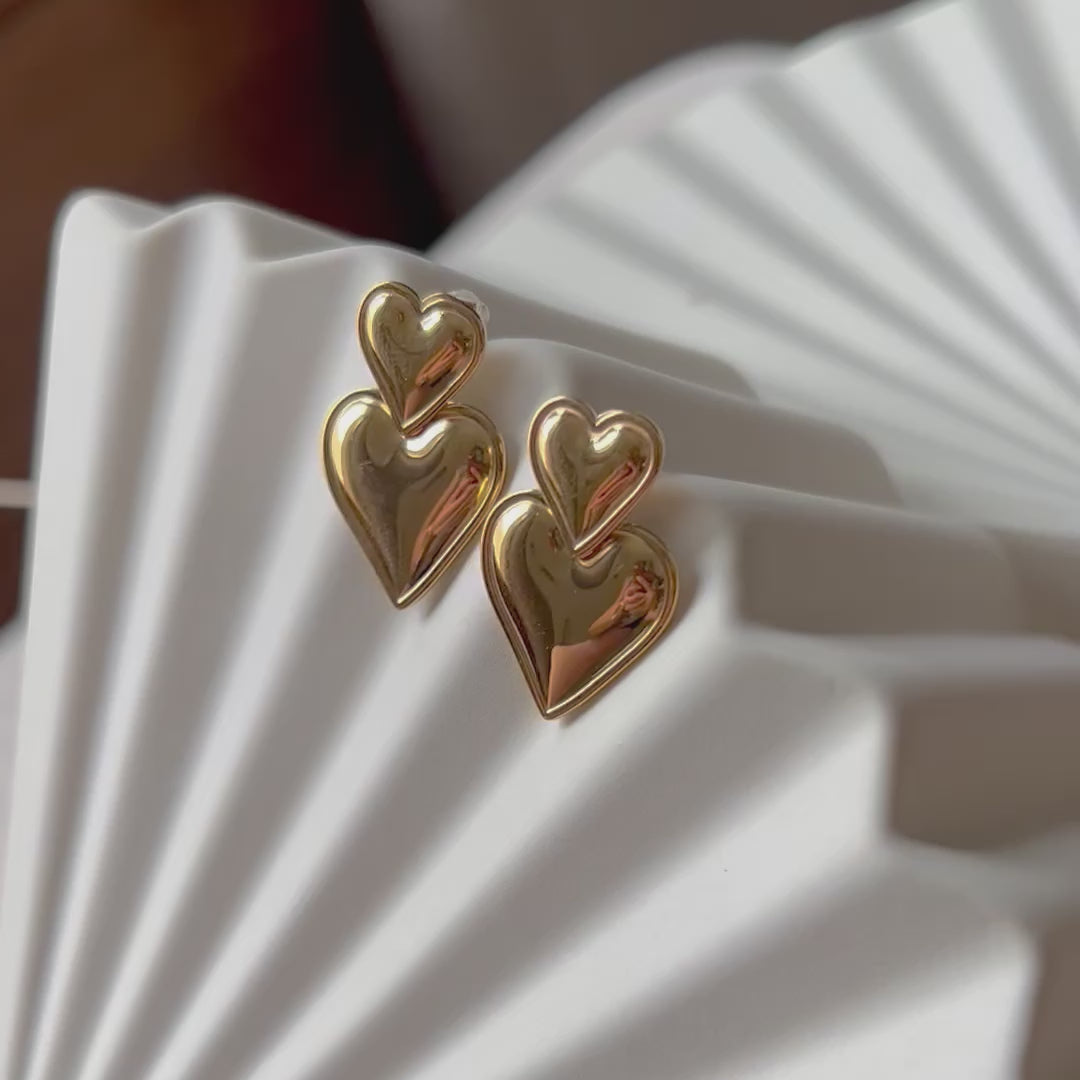Vidéo des boucles d'oreilles "Elle m'a dit" en forme de double cœur, en acier. Posées sur un fond beige texturé. 
