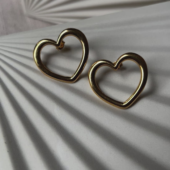 Vidéo des boucles d'oreilles Douce force dorées, grand cœur en acier, posées sur un support beige texturé.