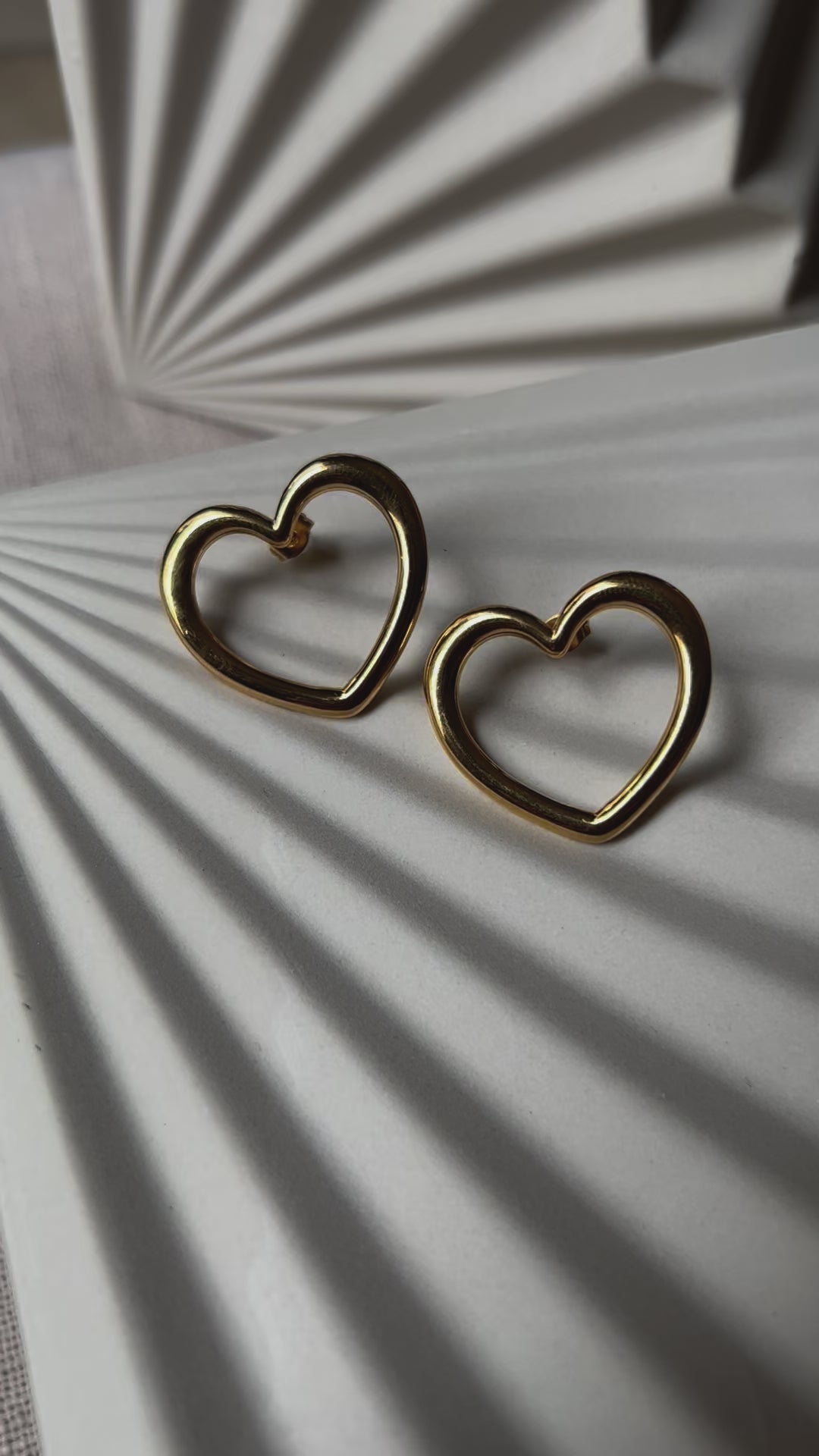 Vidéo des boucles d'oreilles Douce force dorées, grand cœur en acier, posées sur un support beige texturé.