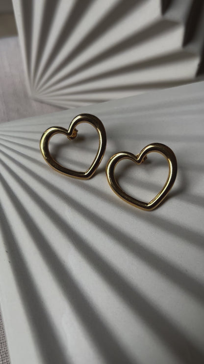 Vidéo des boucles d'oreilles Douce force dorées, grand cœur en acier, posées sur un support beige texturé.