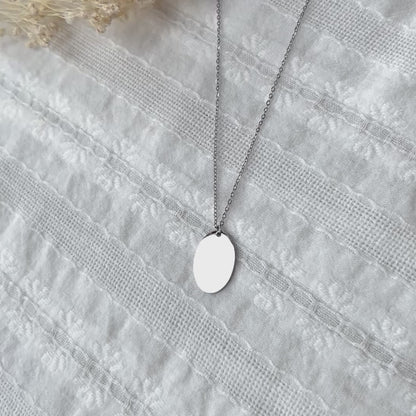 Vidéo du collier "Tendresse", pendentif ovale personnalisable en acier inoxydable argenté, présenté sur un fond beige texturé.