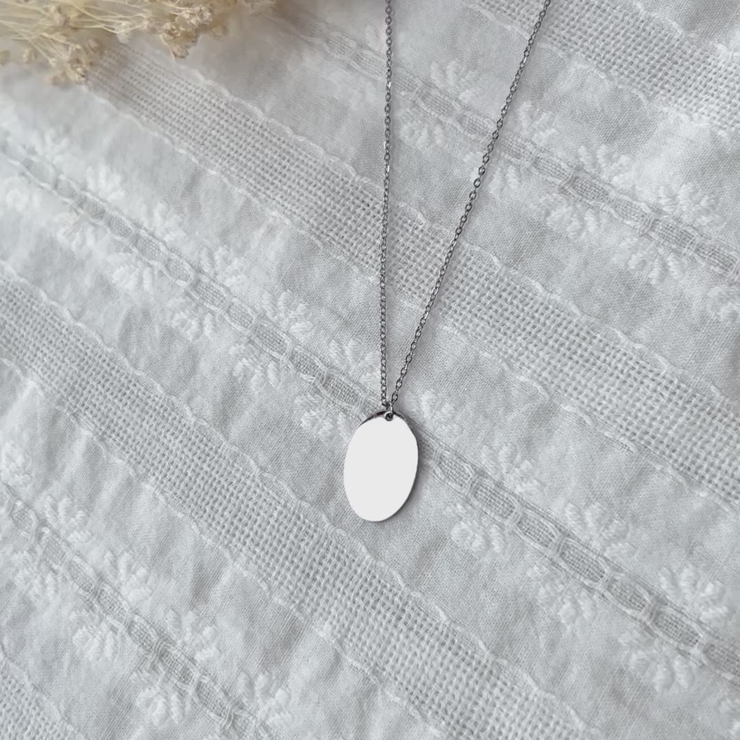 Vidéo du collier "Tendresse", pendentif ovale personnalisable en acier inoxydable argenté, présenté sur un fond beige texturé.