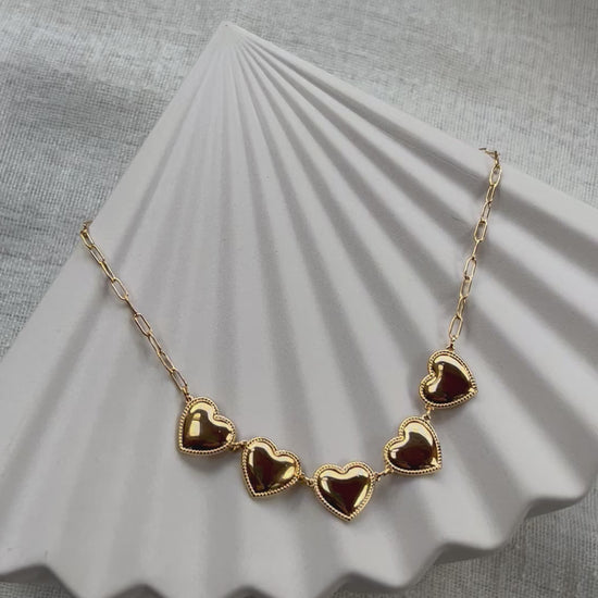 Vidéo du collier "Je suis là" de Visée, composé de cœurs personnalisables en acier doré, posé sur un fond beige texturé.