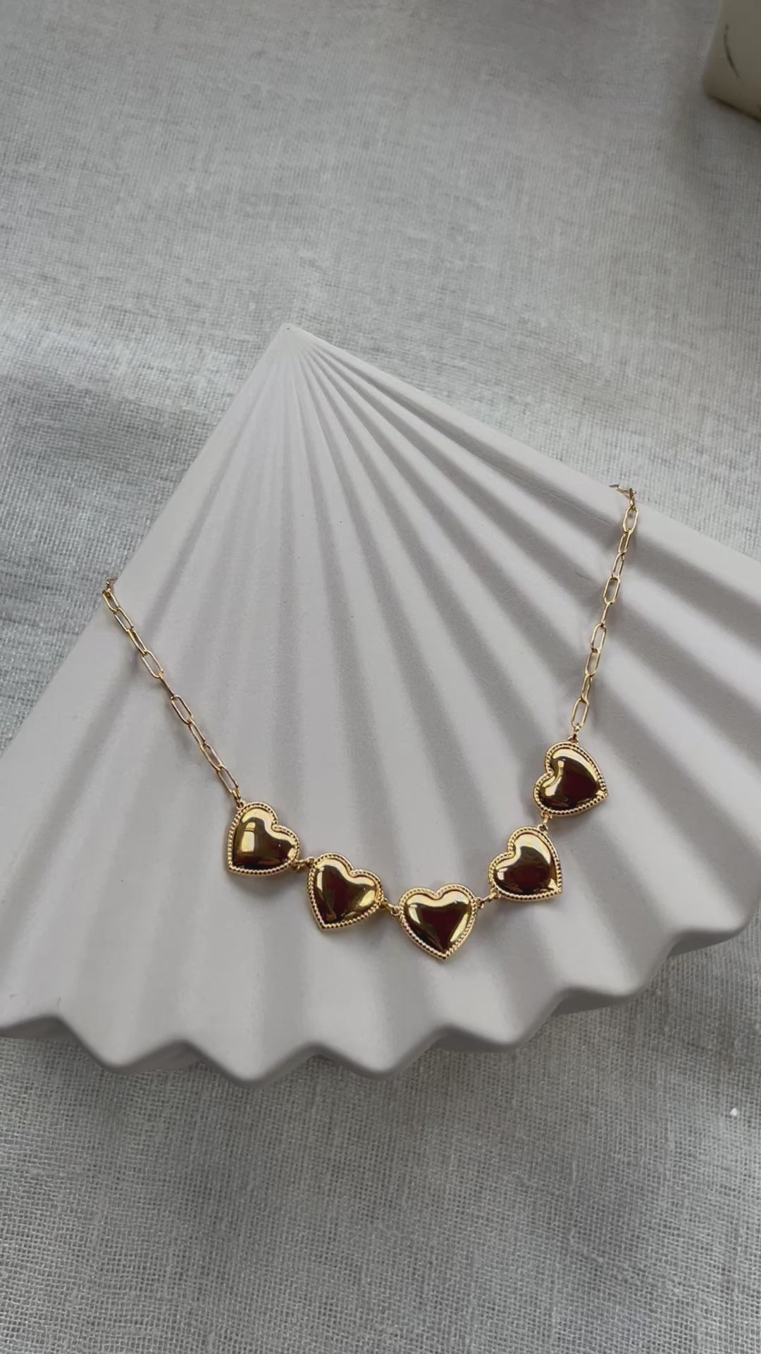 Vidéo du collier "Je suis là" de Visée, composé de cœurs personnalisables en acier doré, posé sur un fond beige texturé.