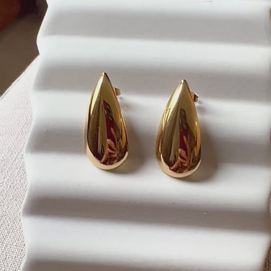 Vidéo de boucles d'oreilles Larmes assumées en  acier doré, en forme de goutte, posées sur un support beige texturé.