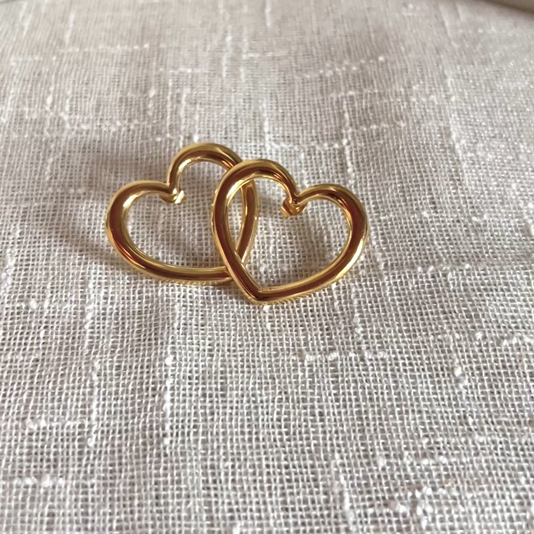 Vidéo des boucles d'oreilles Douce force dorées, grand cœur en acier, posées sur un support beige texturé.