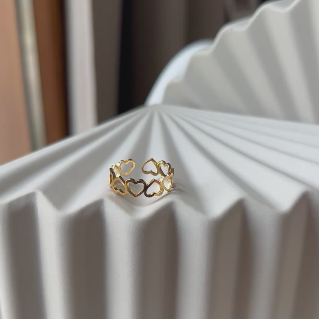 Vidéo de la bague "Unité" réglable en acier doré, composée de cœurs entrelacés, posé sur un support beige texturé.