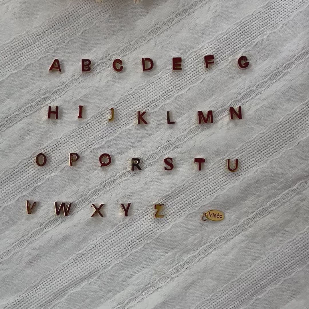 Vidéo de l'ensemble des pendentifs lettres dorées en acier inoxydable, soigneusement alignés  sur un tissu beige texturé.