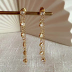 Vidéo boucles d'oreilles dorées Eclat, pendantes et serties de strass brillants, en acier. Photographiées en gros plan sur un support beige texturé.