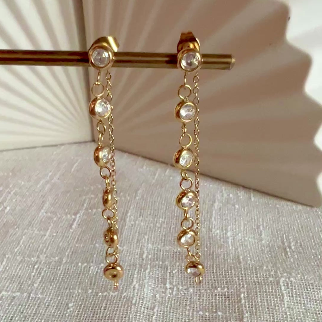 Vidéo boucles d'oreilles dorées Eclat, pendantes et serties de strass brillants, en acier. Photographiées en gros plan sur un support beige texturé.