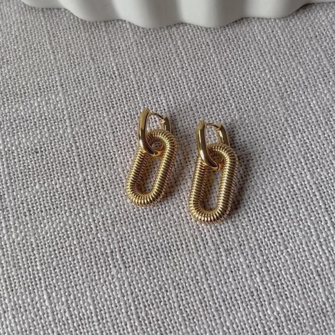 Vidéo boucles d'oreilles "J'existe" en acier doré, au design modulable, posées sur fond beige texturé.
