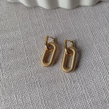 Vidéo boucles d'oreilles "J'existe" en acier doré, au design modulable, posées sur fond beige texturé.