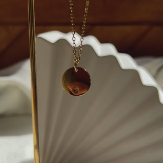 Vidéo du collier personnalisable "Lumineuse", orné d'une médaille ronde brillante en acier doré, posé sur un fond beige texturé.