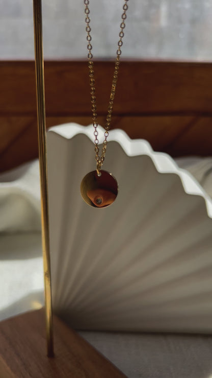 Vidéo du collier personnalisable "Lumineuse", orné d'une médaille ronde brillante en acier doré, posé sur un fond beige texturé.
