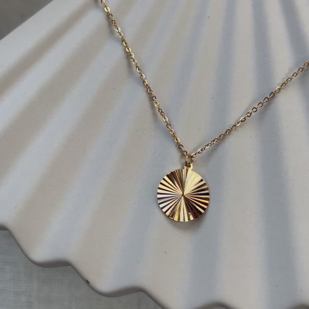 Vidéo du collier "Energie", pendentif rond strié personnalisable en acier doré, posé sur une structure beige.
