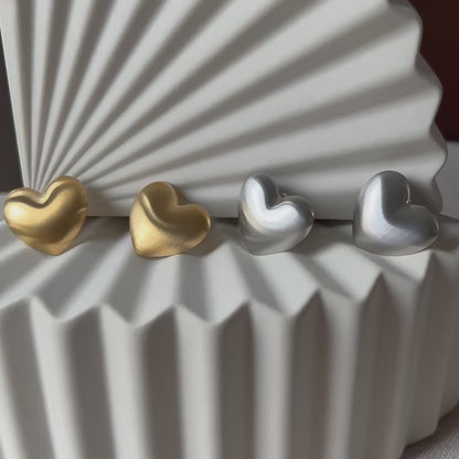 Vidéo des boucles d'oreilles "S'aimer", en maxi cœurs dorés et argentés mat, en acier Inoxydable, posées sur un fond beige texturé.
