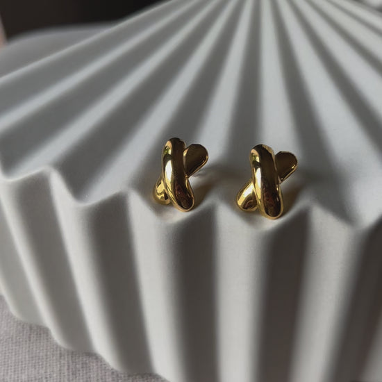 Vidéo des boucles d'oreilles "Révèle toi", au design croix en acier inoxydable doré, photographiées sur un fond beige texturé.