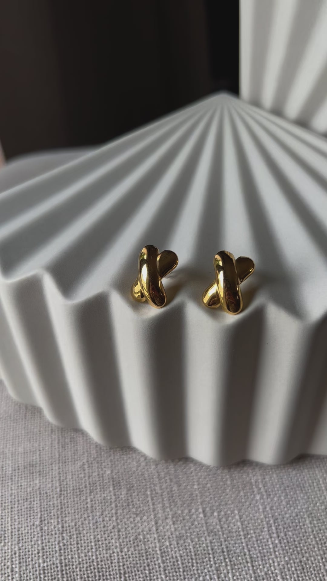 Vidéo des boucles d'oreilles "Révèle toi", au design croix en acier inoxydable doré, photographiées sur un fond beige texturé.