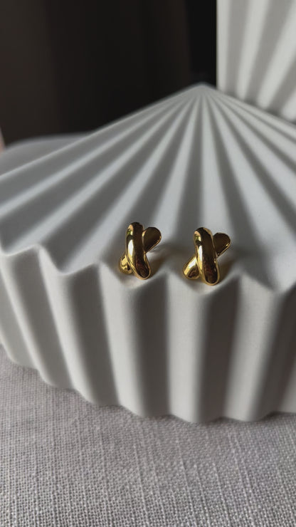 Vidéo des boucles d'oreilles "Révèle toi", au design croix en acier inoxydable doré, photographiées sur un fond beige texturé.