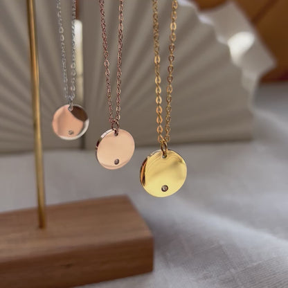 Vidéo du collier personnalisable "Lumineuse", orné d'une médaille ronde brillante en acier, posé sur un fond beige texturé.