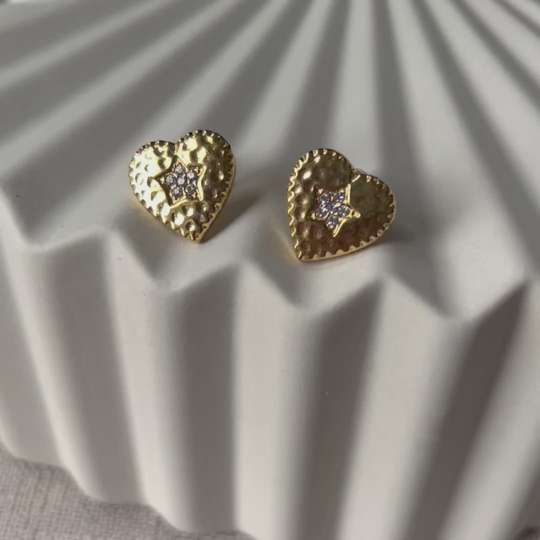 Vidéo des boucles d'oreilles Promesse, en forme de cœur ornées d'une étoile, en acier inoxydable doré, posées sur un support beige texturé.