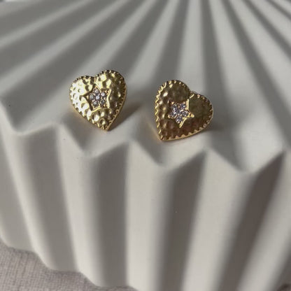 Vidéo des boucles d'oreilles Promesse, en forme de cœur ornées d'une étoile, en acier inoxydable doré, posées sur un support beige texturé.