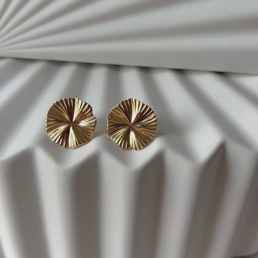 Vidéo boucles d'oreilles "Ma raison" en acier doré, au motif floral, photographiées sur un fond plissé beige texturé.