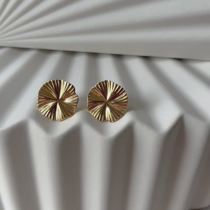 Vidéo boucles d'oreilles "Ma raison" en acier doré, au motif floral, photographiées sur un fond plissé beige texturé.